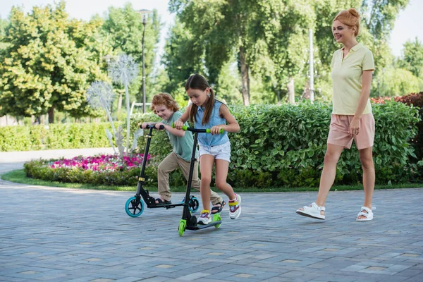 Mujer feliz caminando cerca de los niños montando patinete scooters en el parque - foto de stock