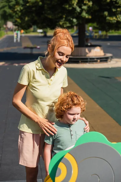 Happy redhead woman hugging shoulders of son on playground — Stock Photo