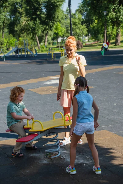 Pleased woman standing near children and seesaw in park — Stock Photo