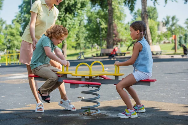 Bambini felici cavalcando altalena vicino alla madre nel parco — Foto stock