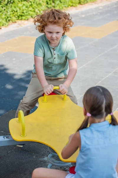 Curly boy riding seesaw with blurred sister outdoors — Stock Photo