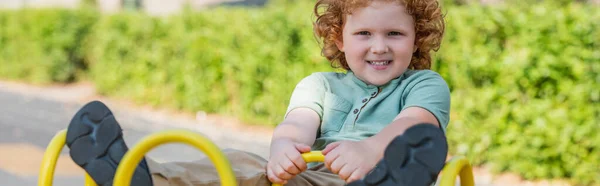 Excited redhead kid looking at camera while riding seesaw in park, banner — Stock Photo