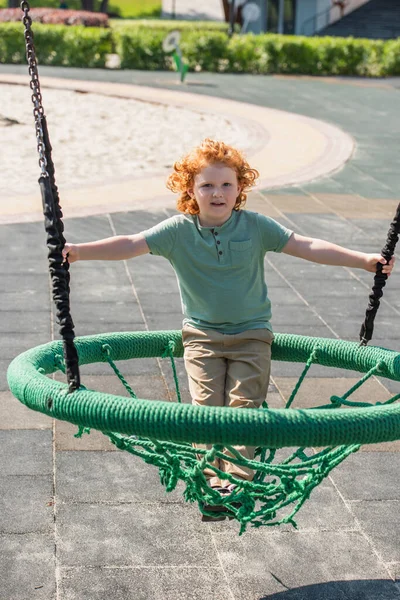 Lockiger Junge schaut in die Kamera, während er Spaß in der Hängematte auf dem Spielplatz hat — Stockfoto