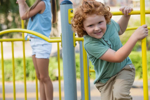 Excited boy looking at camera while playing near blurred sister on playground — Stock Photo