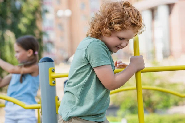 Riccio ragazzo giocare vicino sfocato ragazza su parco giochi — Foto stock