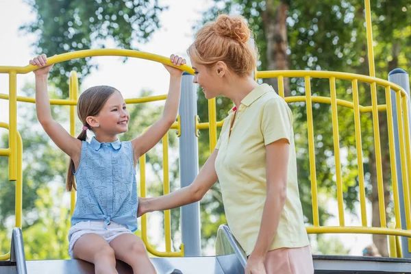 Femme regardant fille heureuse s'amuser sur la glissière dans le parc d'attractions — Photo de stock