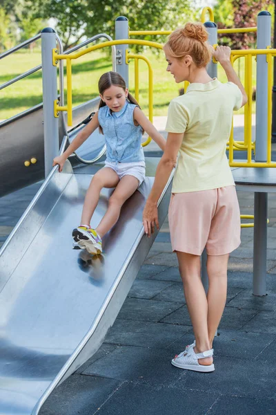 Vue pleine longueur de la femme debout près de la fille sur la glissière dans le parc d'attractions — Photo de stock