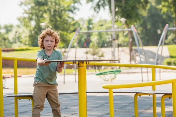 Redhead boy having fun on carousel in park and looking at camera — Stock Photo