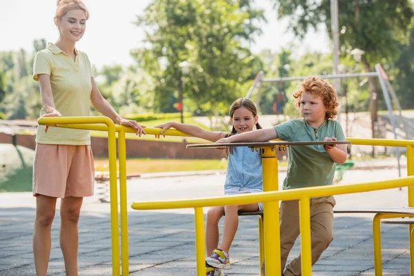 Mujer alegre de pie cerca de los niños en carrusel en el parque - foto de stock