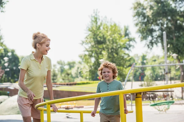 Donna rossa allegra sorridente vicino a figlio su giostra in parco — Foto stock