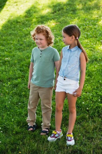 Full length view of brother and sister in summer clothes standing on green lawn — Stock Photo