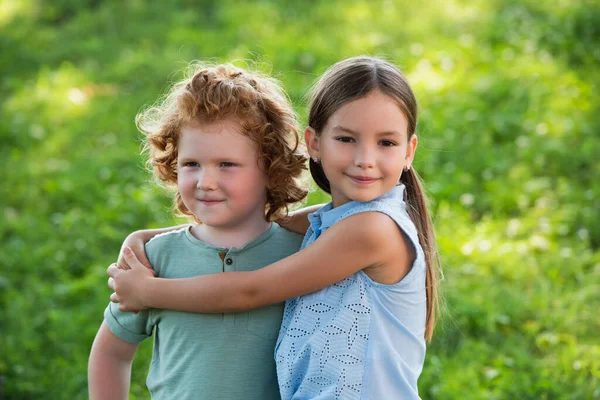 Sonriente chica abrazando hermano y mirando a la cámara al aire libre - foto de stock