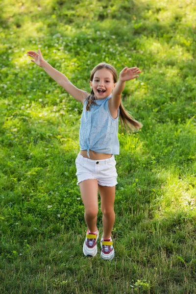 Full length view of excited girl in shorts waving hands while running on green lawn — Stock Photo