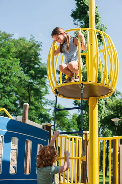 Boy climbing on rope ladder under sister on high tower in amusement park — Stock Photo
