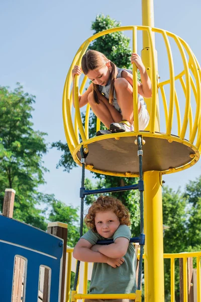 Boy on rope ladder looking at camera near sister on high tower in amusement park — Stock Photo