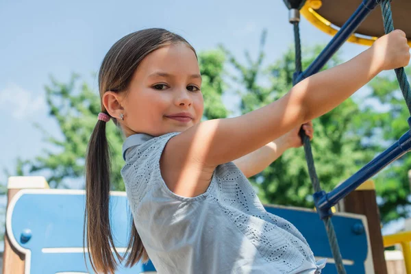 Fille souriant à la caméra tout en grimpant sur l'échelle de corde sur aire de jeux — Photo de stock