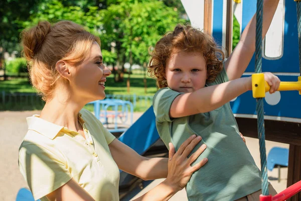 Sonriente pelirroja mujer de apoyo hijo subir en cuerda escalera en parque infantil - foto de stock