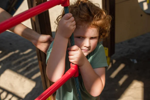 Ricci ragazzo vicino corda scala e madri mano nel parco divertimenti — Foto stock