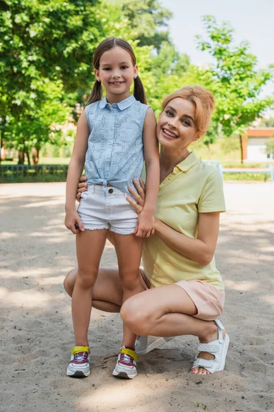 Full length view of girl in summer clothes looking at camera near happy mother in park — Stock Photo
