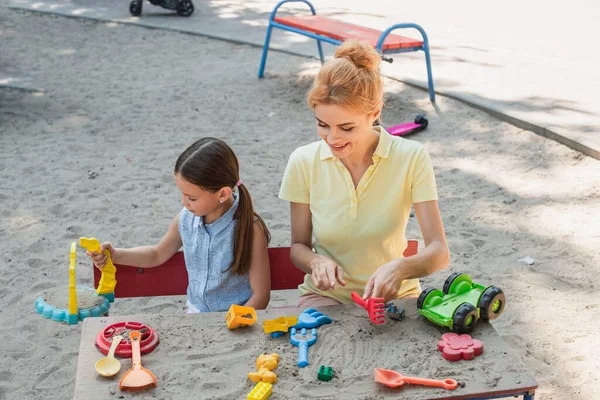 Femme souriante avec fille jouant avec des jouets et du sable à l'extérieur — Photo de stock