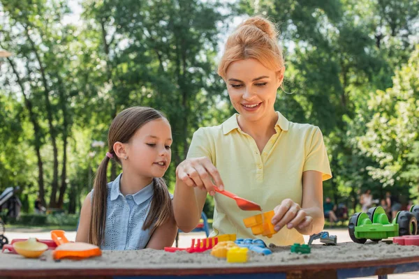 Donna sorridente che gioca con pala giocattolo e sabbia vicino figlia — Foto stock