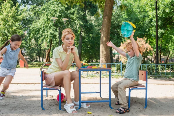 Verärgerte Frau sitzt neben glücklichen Kindern und amüsiert sich auf Spielplatz — Stockfoto