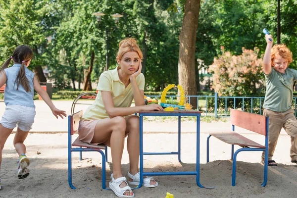 Sad woman looking at camera near children having fun on playground — Stock Photo