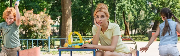 Donna triste guardando la macchina fotografica vicino ai bambini che corrono sul parco giochi, striscione — Foto stock