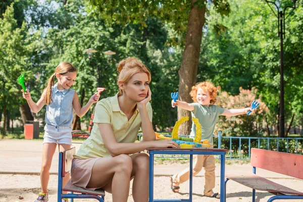 Mujer triste sentada y mirando lejos cerca de niños alegres jugando con rastrillos de juguete - foto de stock