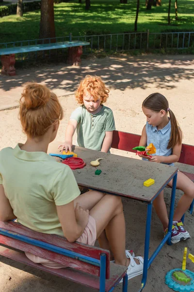 Niños jugando con juguetes y arena cerca de la madre en el patio de recreo - foto de stock