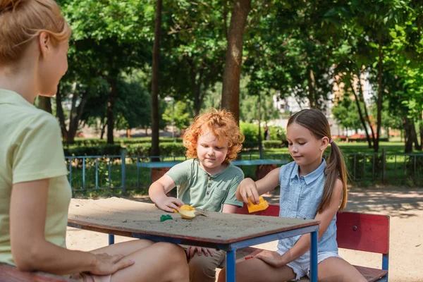 Niños jugando con juguetes y arena mientras están sentados cerca de la madre en el patio de recreo - foto de stock