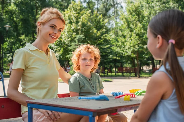 Happy woman sitting near kids and toy rakes on playground — Stock Photo