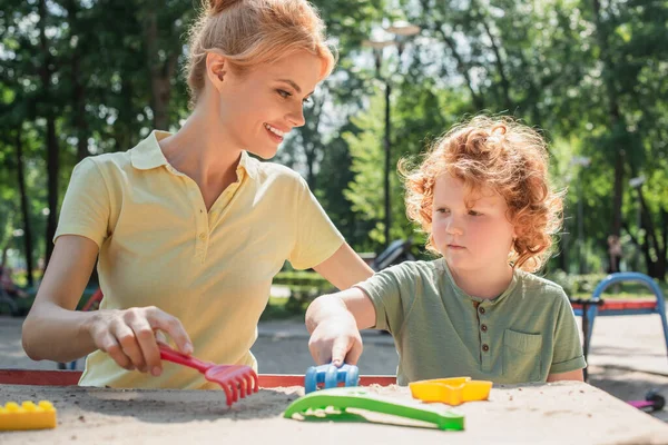 Ricci ragazzo giocare con giocattolo rastrelli e sabbia vicino allegro madre — Foto stock