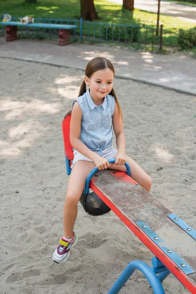 Smiling girl in shorts riding seesaw in summer park — Stock Photo