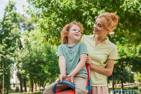 Счастливая рыжая женщина улыбается рядом с сыном катаясь на качелях по детской площадке — стоковое фото