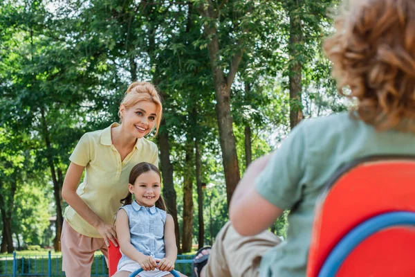 Femme souriant près des enfants équitation balançoire sur le premier plan flou — Photo de stock