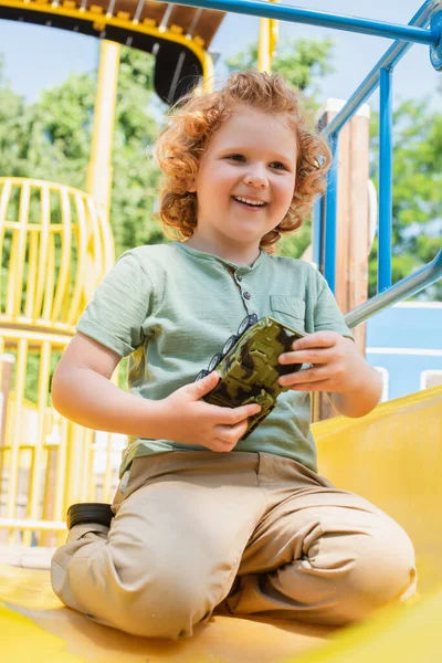 Alegre chico sosteniendo juguete vehículo blindado en diapositiva en parque de atracciones - foto de stock