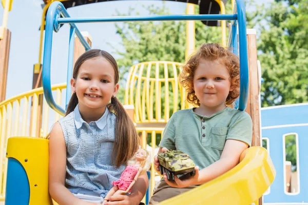 Glückliche Kinder mit Puppe und Spielzeug Panzerfahrzeug Blick auf Kamera in Freizeitpark — Stockfoto