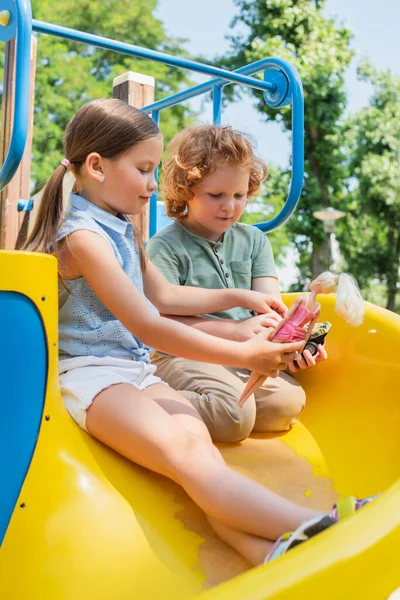Niños jugando con muñeca y coche de juguete mientras están sentados en la diapositiva - foto de stock