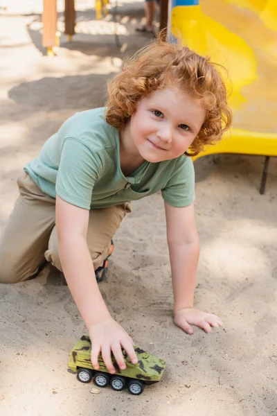Niño rizado mirando a la cámara mientras juega en la arena con vehículo blindado de juguete - foto de stock