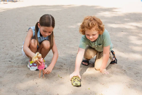 Vista completa de los niños jugando con la muñeca y el vehículo blindado en la arena al aire libre - foto de stock