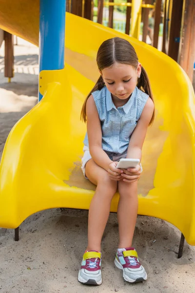 Vue pleine longueur de fille en vêtements d'été jouer sur smartphone tout en étant assis sur l'aire de jeux — Photo de stock