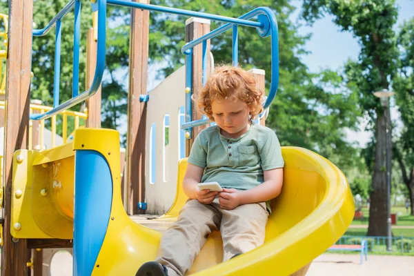 Redhead boy sitting on slide in amusement park and playing on smartphone — Stock Photo