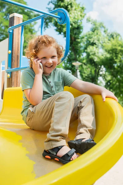 Alegre chico hablando en smartphone en diapositiva en parque de atracciones - foto de stock