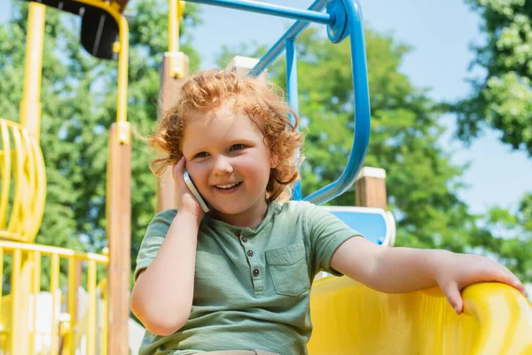 Niño sonriente llamando en el teléfono móvil en la diapositiva en el parque de atracciones - foto de stock