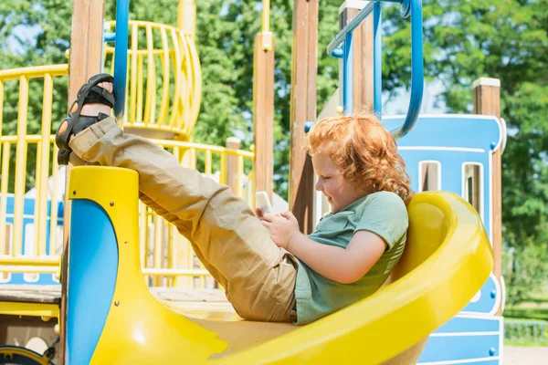 Vista laterale del ragazzo allegro seduto sullo scivolo e giocare sul telefono cellulare — Foto stock