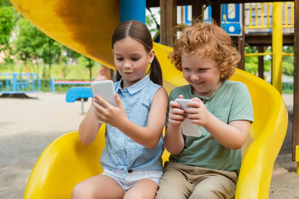 Aufgeregtes Geschwisterpaar sitzt auf Rutsche und spielt auf Smartphone — Stockfoto