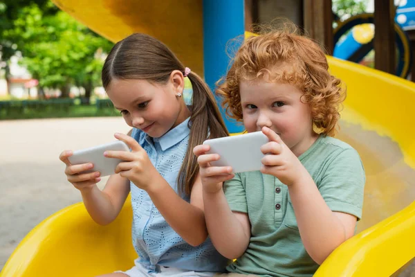 Niño con el teléfono móvil mirando a la cámara cerca de la hermana en el patio de recreo - foto de stock