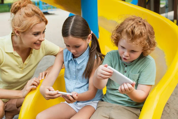 Fröhliche Frau in der Nähe von Kindern, die auf Spielplatz mit Smartphones spielen — Stockfoto