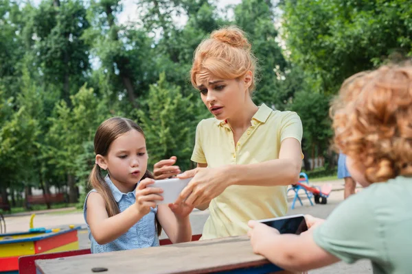 Donna dispiaciuta prendendo smartphone da figlia mentre seduto sul parco giochi — Foto stock